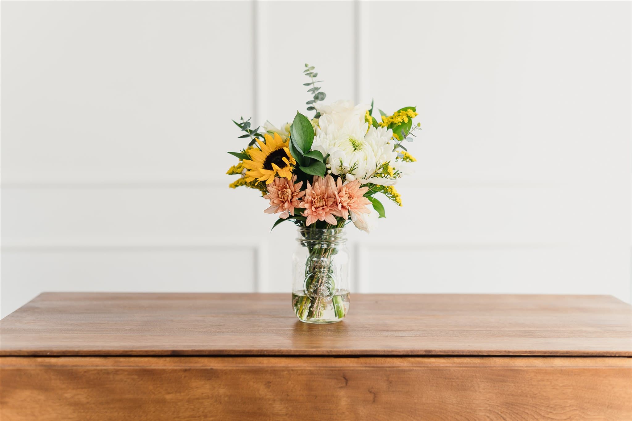 Bouquet BeauBlooms avec tournesol et chrysanthèmes dans un pot en verre, posé sur une table en bois